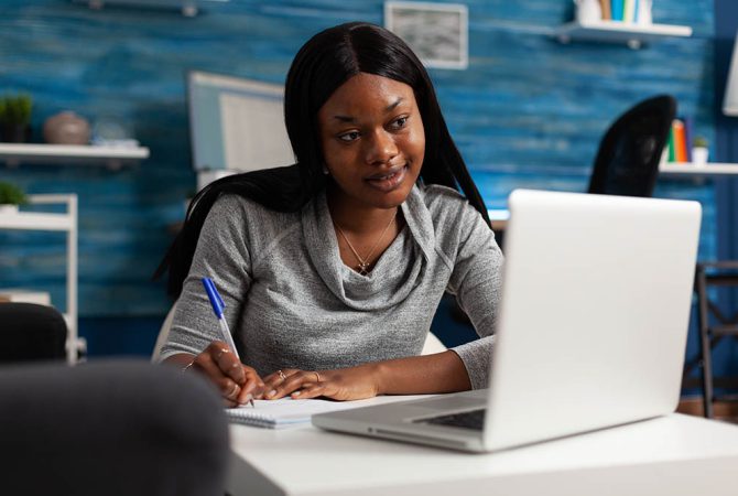 Young person taking notes on textbook paper with pen, looking at modern laptop. Woman writing information on notebook files and doing remote work. Adult working from home on business.