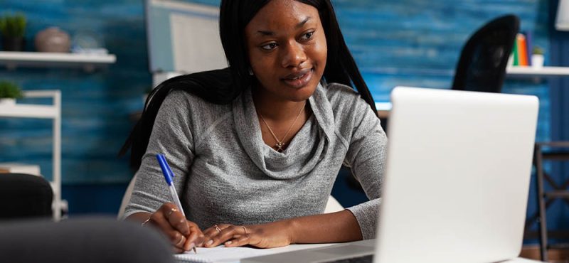 Young person taking notes on textbook paper with pen, looking at modern laptop. Woman writing information on notebook files and doing remote work. Adult working from home on business.